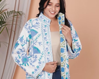May include: White jacket with blue floral embroidery. The jacket has a stand-up collar and long sleeves. The floral design is in shades of blue and green. The jacket is open in the front and is worn over a light-colored top and blue jeans.