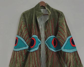 May include: Olive green velvet jacket with a unique eye design. The jacket features large, turquoise and red eye appliqués on the sleeves. The interior lining is striped. The jacket is hanging on a wooden hanger.