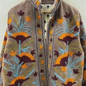 Puede incluir: Chaqueta marrón con intrincados bordados florales en naranja, burdeos y azul. La chaqueta tiene un cuello alto y un forro estampado visible en el escote. El bordado crea un diseño texturizado en la parte delantera.