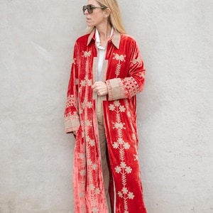 May include: A long, red velvet coat with floral embroidery. The coat features a collar and cuffs with a tan trim. The person is wearing tan pants and white sneakers. The coat is a statement piece.