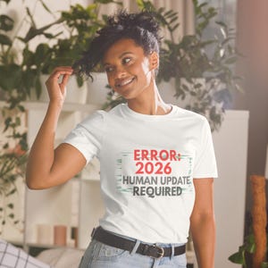 May include: White t-shirt with the text "ERROR 2026 HUMAN UPDATE REQUIRED" in red and gray. The shirt has a crew neck and short sleeves. The person is wearing blue jeans and a black belt.