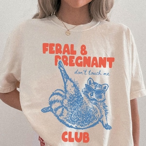 May include: An ivory short-sleeve t-shirt featuring a blue graphic of a raccoon wearing sunglasses with one leg raised. The text reads FERAL & PREGNANT in orange and don't touch me in blue, with the word CLUB in orange at the bottom of the graphic.
