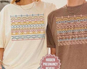 Może przedstawiać: Dwie koszulki z krótkim rękawem, jedna w kolorze kości słoniowej, a druga w kolorze espresso, każda z prostokątnym nadrukiem graficznym przedstawiającym poziome rzędy geometrycznych wzorów w stonowanych odcieniach niebieskiego, różu i złota.