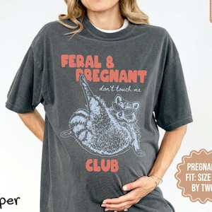 May include: A charcoal gray short-sleeve t-shirt featuring a graphic of a raccoon wearing sunglasses with the text FERAL & PREGNANT don't touch me CLUB in coral lettering. The shirt has a crew neckline and a relaxed fit with a vintage-style wash.