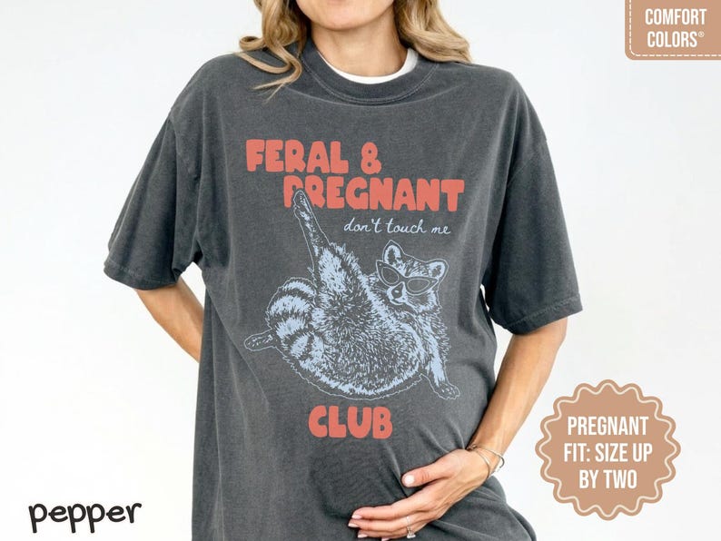 May include: A charcoal gray short-sleeve t-shirt featuring a graphic of a raccoon wearing sunglasses with the text FERAL & PREGNANT don't touch me CLUB in coral lettering. The shirt has a crew neckline and a relaxed fit with a vintage-style wash.