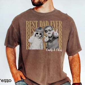 Pu&ograve; includere: T-shirt color espresso con grafica dorata "BEST DAD EVER". Il design include due foto in bianco e nero di bambini con occhiali da sole. I nomi "Emily & Chris" sono stampati sotto le foto.