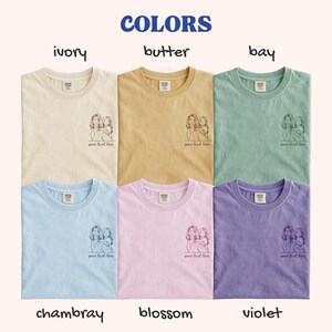 Puede incluir: Seis camisetas en varios colores: marfil, mantequilla, bah&iacute;a, chambray, flor y violeta. Cada camiseta tiene cuello redondo y un peque&ntilde;o gr&aacute;fico de dos figuras con el texto "your text here" debajo. La palabra "COLORS" est&aacute; en la parte superior.
