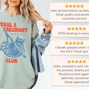 May include: A sage green t-shirt featuring a blue graphic of a raccoon wearing sunglasses with the red text FERAL & PREGNANT CLUB and the blue text don't touch me. The shirt includes a Comfort Colors label and is shown with customer reviews.