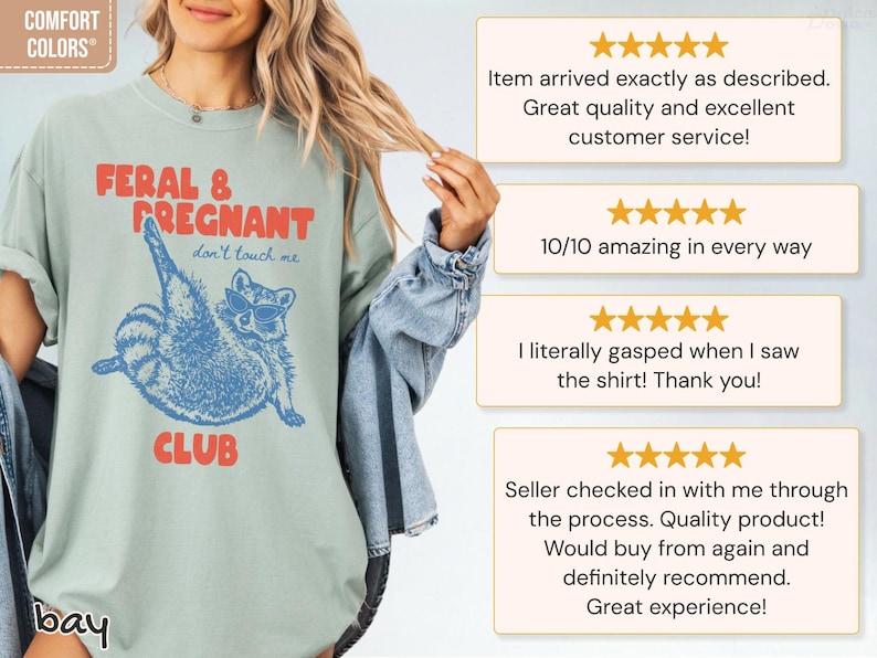 May include: A sage green t-shirt featuring a blue graphic of a raccoon wearing sunglasses with the red text FERAL & PREGNANT CLUB and the blue text don't touch me. The shirt includes a Comfort Colors label and is shown with customer reviews.
