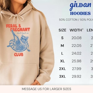 May include: A beige hooded sweatshirt featuring a blue graphic of a raccoon wearing sunglasses with the red text FERAL & PREGNANT CLUB and don't touch me. A size chart displays widths and lengths in inches for sizes S through 3XL, noting 50% cotton and 50% polyester.