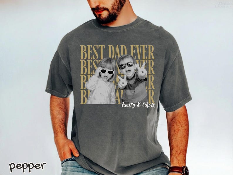 Pu&ograve; includere: T-shirt grigio scuro con la scritta dorata "BEST DAD EVER". La maglietta presenta una foto in bianco e nero di due bambini con occhiali da sole. I nomi "Emily & Chris" sono stampati sotto.