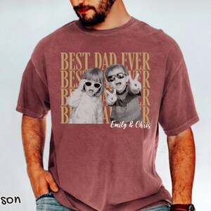 Pu&ograve; includere: T-shirt color cremisi con la scritta "BEST DAD EVER" in oro. La maglietta presenta una foto in bianco e nero di due bambini con occhiali da sole. I nomi "Emily & Chris" sono stampati sotto.