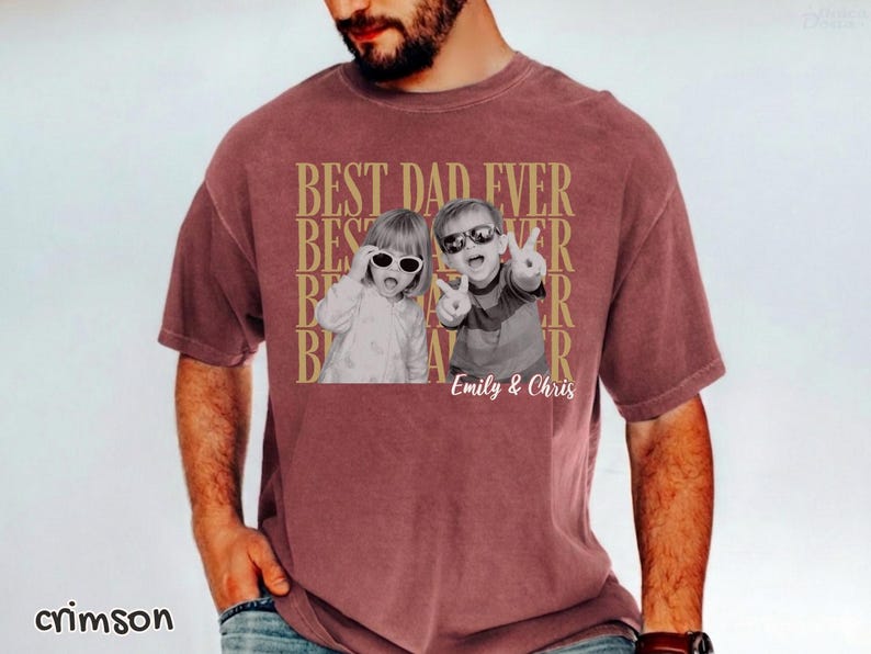Pu&ograve; includere: T-shirt color cremisi con la scritta "BEST DAD EVER" in oro. La maglietta presenta una foto in bianco e nero di due bambini con occhiali da sole. I nomi "Emily & Chris" sono stampati sotto.