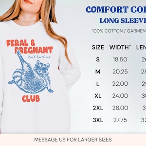 May include: A white long-sleeve shirt featuring a blue graphic of a raccoon wearing sunglasses with the red text FERAL & PREGNANT CLUB and don't touch me. A size chart lists widths and lengths in inches for sizes S through 3XL, noting 100% cotton.