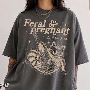 May include: A charcoal gray short-sleeve t-shirt featuring a graphic of a raccoon wearing sunglasses with one leg raised. The text reads Feral & pregnant don't touch me in a light beige font, accented by small star shapes across the chest area.