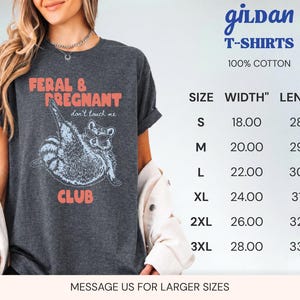 May include: A dark gray short-sleeve t-shirt featuring a graphic of a raccoon wearing sunglasses with the text FERAL & PREGNANT don't touch me CLUB. A size chart is visible with width and length measurements in inches for sizes S to 3XL.