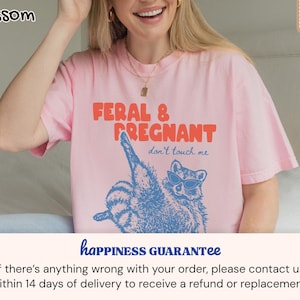 May include: A light pink short-sleeve t-shirt featuring a blue graphic of a raccoon wearing sunglasses. The text reads FERAL & PREGNANT in orange and don't touch me in blue. The shirt has a crew neckline and a relaxed fit, made of cotton fabric.