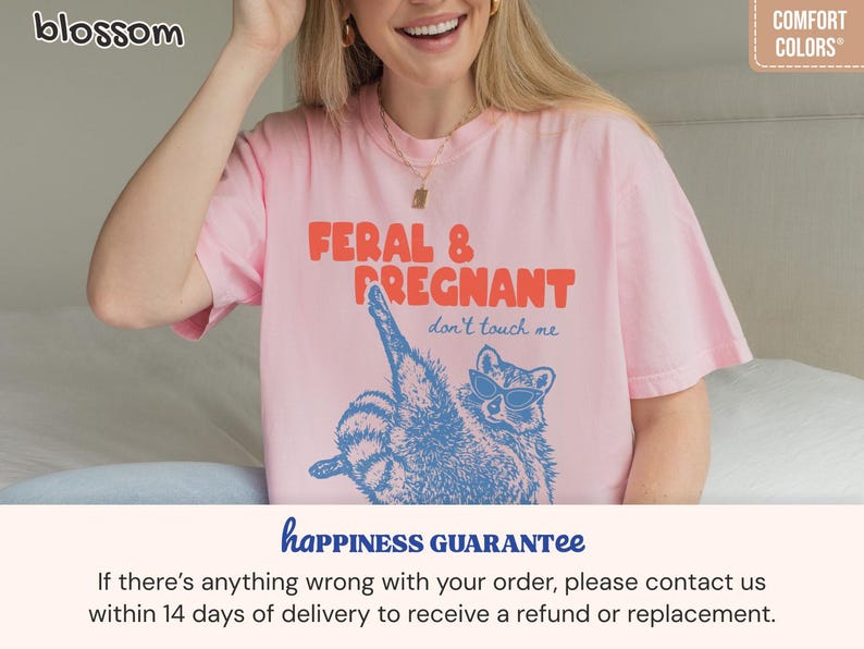 May include: A light pink short-sleeve t-shirt featuring a blue graphic of a raccoon wearing sunglasses. The text reads FERAL & PREGNANT in orange and don't touch me in blue. The shirt has a crew neckline and a relaxed fit, made of cotton fabric.