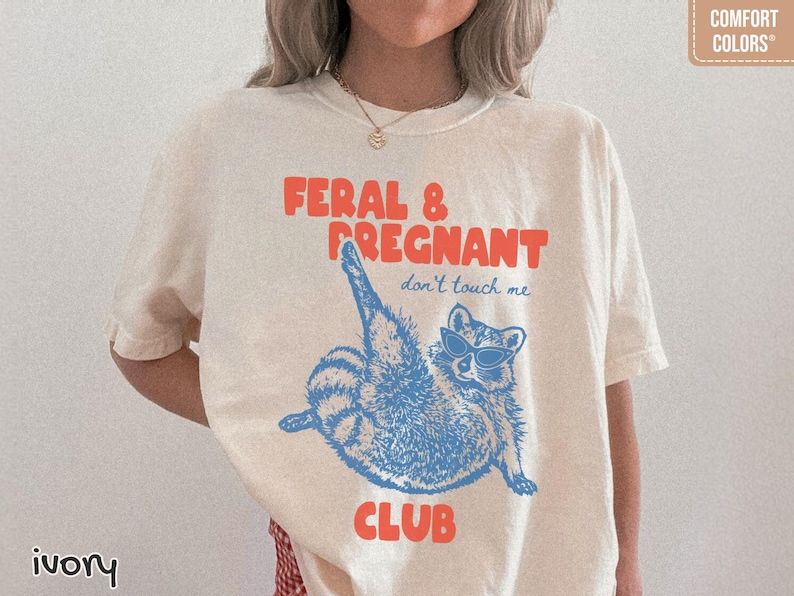 May include: An ivory short-sleeve t-shirt featuring a blue graphic of a raccoon wearing sunglasses with one leg raised. The text reads FERAL & PREGNANT in orange and don't touch me in blue, with the word CLUB in orange at the bottom of the graphic.