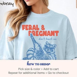 May include: A light blue t-shirt featuring the red text FERAL & PREGNANT and the blue text don't touch me. Below the text is a blue graphic illustration of a raccoon wearing sunglasses, with one leg raised. The shirt has a crew neckline.