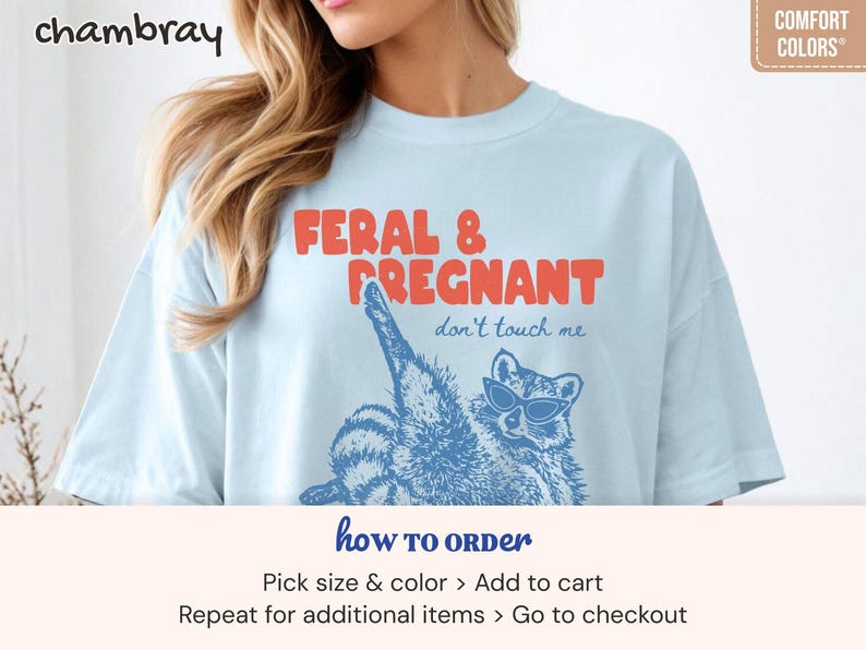 May include: A light blue t-shirt featuring the red text FERAL & PREGNANT and the blue text don't touch me. Below the text is a blue graphic illustration of a raccoon wearing sunglasses, with one leg raised. The shirt has a crew neckline.