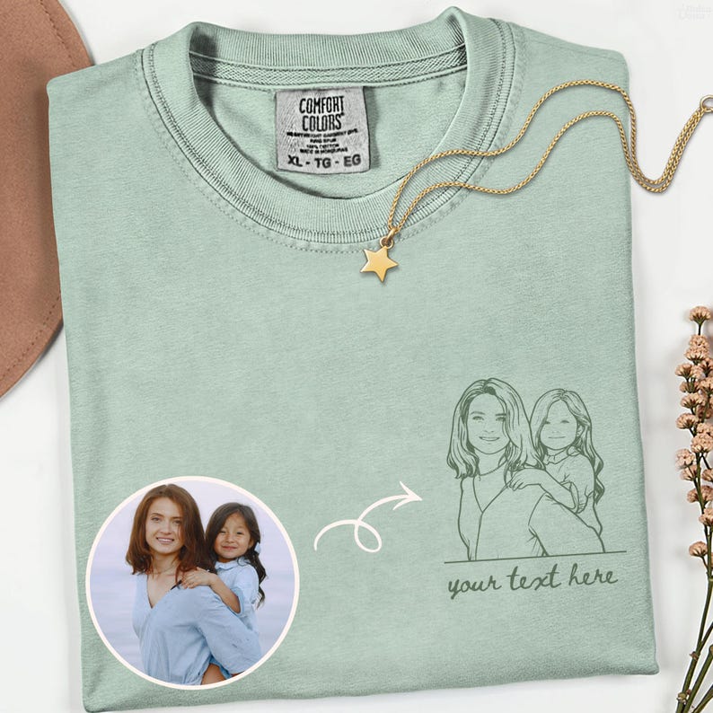 Puede incluir: Una camiseta Comfort Colors XL verde claro con un collar con colgante de estrella dorada. La camiseta presenta un boceto de una madre y un ni&ntilde;o, una foto de la misma pareja y el texto "your text here".