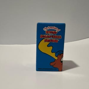 Può includere: Una scatola rettangolare blu con il titolo The Adventures of Rocky and Bullwinkle and Friends in caratteri rossi e gialli. Un'illustrazione parziale di un alce marrone con un grande palco giallo è visibile nell'angolo in basso a destra.