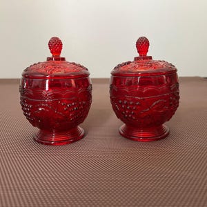 May include: Two matching ruby red glass jars with lids, each featuring a raised embossed pattern of grape clusters and leaves. The jars have a rounded body, a pedestal base, and a small knob on the lid shaped like a cluster of grapes.