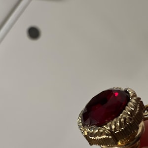 May include: A gold-toned metal ring featuring a large, faceted, deep red gemstone set in a textured, rope-style border. The circular stone is polished and reflects light, while the metal setting displays a detailed, ridged pattern around the edge.