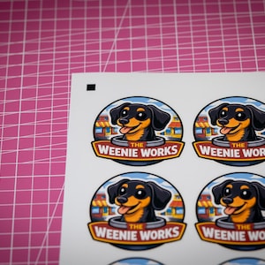 May include: Sheet of stickers featuring a cartoon dachshund dog with a smiling expression. The dog is black and tan, set against a blue and white background. The text "The Weenie Works" is displayed in a banner below the dog.