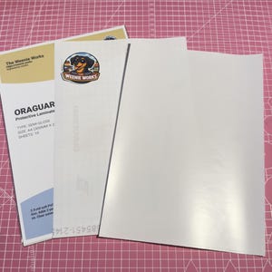 May include: A stack of white protective laminate sheets with the text "ORAGUAR Protective Laminate" and "The Weenie Works" printed on the packaging. The packaging includes a logo of a dachshund dog.