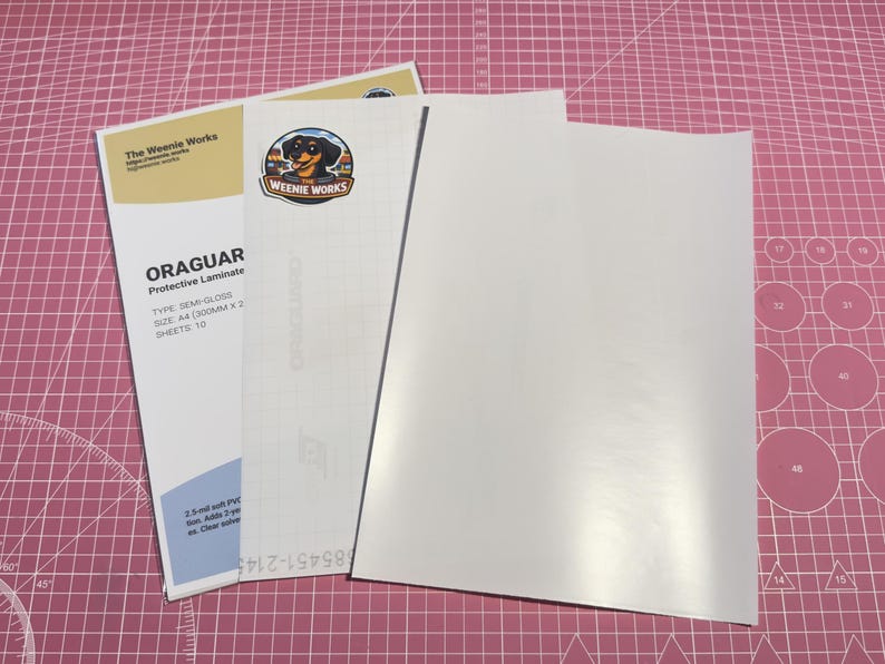May include: A stack of white protective laminate sheets with the text "ORAGUAR Protective Laminate" and "The Weenie Works" printed on the packaging. The packaging includes a logo of a dachshund dog.