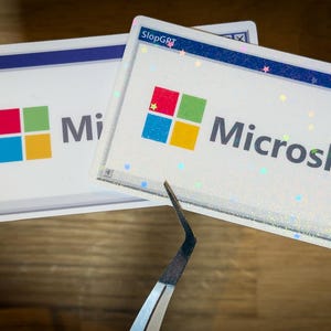 Puede incluir: Dos pegatinas blancas con un logotipo inspirado en Microsoft, con un diseño cuadrado colorido. El texto "Microslop" se muestra en una pegatina y "Mi" en la otra. Las pegatinas tienen un borde azul y están sobre una superficie de madera.