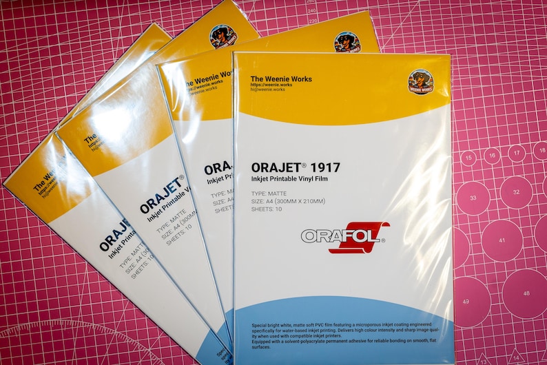 May include: Several packages of ORAJET 1917 inkjet printable vinyl film are fanned out on a pink cutting mat. The packages have a white and yellow design with the text "The Weenie Works" and "ORAFOL". Each sheet is A4 size (300mm x 210mm).