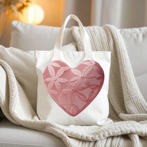 Puede incluir: Un bolso tote blanco con un gran gráfico de corazón centrado. El corazón es de un tono rosa suave y muestra un intrincado patrón floral similar al encaje, con pétalos interconectados y una textura de malla fina en toda su superficie.