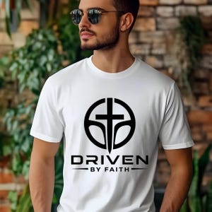 Puede incluir: Camiseta blanca con un diseño gráfico negro que presenta una cruz dentro de un círculo y las palabras "DRIVEN BY FAITH". La camiseta está hecha de un material suave y es adecuada para uso casual.