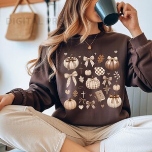 Puede incluir: Una sudadera de manga larga en color marrón oscuro con un patrón repetitivo de calabazas, lazos, corazones, cruces y un pequeño libro en tonos beige y blanco. El estampado gráfico está centrado en el pecho con iconos de temática otoñal.