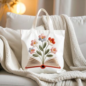 Puede incluir: Bolso tote blanco con un diseño floral bordado y un libro abierto. Las flores son rosas, azules y naranjas, con hojas verdes. El bolso tiene dos asas blancas.