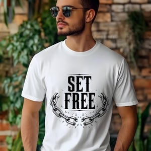 Puede incluir: Una camiseta blanca de manga corta con un estampado gráfico negro en el pecho. El diseño muestra el texto SET FREE en una fuente serif negrita, colocado sobre una ilustración de una cadena de metal rota con eslabones separados en el centro.