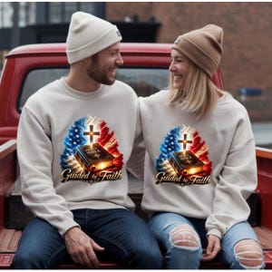 Puede incluir: Sudaderas de color blanquecino con un gráfico que presenta una cruz, una Biblia y el texto "Guided by Faith". El diseño incorpora elementos rojos, blancos y azules. Las sudaderas son usadas por dos personas.