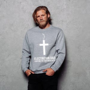 May include: A light gray sweatshirt with the text "ELECTRICIAN DAD LIGHTING THE WAY WITH FAITH" and a white cross design. The cross has a lightning bolt extending from the top. The sweatshirt is long-sleeved.