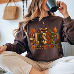 Puede incluir: Una sudadera de manga larga de color marrón oscuro con la palabra BLESSED en letras grandes, texturizadas y con varios patrones. El diseño incluye motivos otoñales como hojas de arce naranjas y rojas, bellotas y lazos de cuadros.