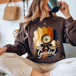 Puede incluir: Una sudadera de manga larga de color marrón oscuro con un estampado gráfico de una brújula dorada vintage sobre un pergamino enrollado atado con una cinta negra. El diseño incluye pinceladas en amarillo, naranja y azul, con la palabra Guided.