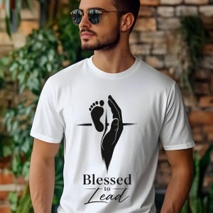 May include: White t-shirt with a black graphic of a hand holding a baby footprint. The text "Blessed to Lead" is below the graphic. The shirt is made of a soft material.
