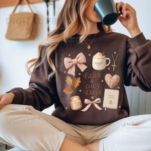 May include: A dark brown long-sleeve sweatshirt featuring a graphic print with the text Faith & cozy Days. The design includes a pink bow, a pumpkin spice mug, a cross necklace, a plaid heart, a prayer journal, a candle, and oak leaves.