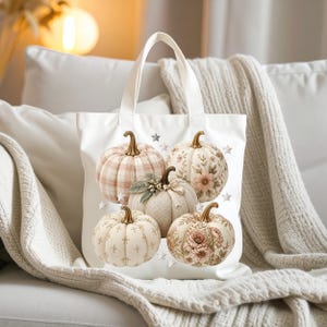 Puede incluir: Un bolso de tela blanco con un diseño estampado de cinco calabazas en varios tonos crema, beige y rosa suave. Las calabazas muestran patrones que incluyen cuadros, motivos florales y diseños de cuadrícula, acentuados por pequeñas estrellas plateadas.