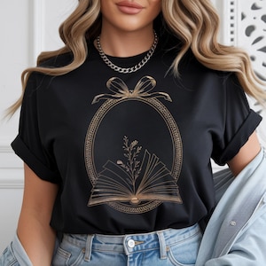 Puede incluir: Camiseta negra con un diseño dorado de marco ovalado, lazo, libro abierto y elementos florales. La camiseta tiene mangas enrolladas y cuello redondo. El diseño está centrado en la parte delantera.