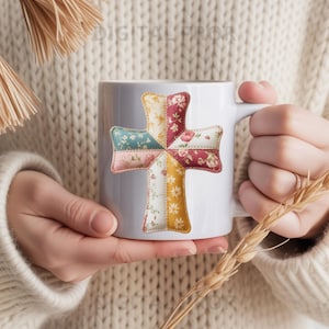 Op de afbeelding: Witte keramische mok met een kleurrijk patchwork kruisontwerp. Het kruis is gemaakt van verschillende bloemen- en patroonstoffen in tinten blauw, roze, geel en rood. De mok wordt vastgehouden door een persoon die een crèmekleurige trui draagt.
