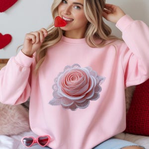 Puede incluir: Sudadera rosa claro con un diseño de rosa grande y detallado con un borde de encaje. La rosa es de color rosa suave con detalles blancos. La sudadera tiene mangas largas y cuello redondo.