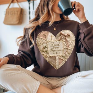 Puede incluir: Una sudadera de manga larga de color marrón oscuro con un gran parche en forma de corazón en el pecho. El parche está compuesto por varias texturas de encaje, tela y bordados florales en beige y crema, con el texto Loved Beyond Measure impreso.